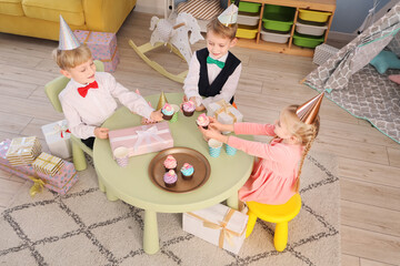 Wall Mural - Cute little happy kids with cupcakes and gift boxes celebrating birthday at table in children's room