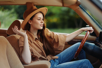 Canvas Print - Stylish young woman driving a vintage car, wearing a brown oversized shirt and hat, exuding confidence and joy in a lush green environment.