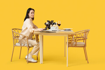 Wall Mural - Young woman sitting at restaurant table on yellow background