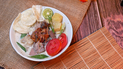 Wall Mural - Top view of a bowl of Soup or Soto Betawi with beef, potato, fresh tomato, lime slices, and fried shallot. Served on bamboo mat and burlap. Space for text available