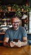 © Maria - Smiling man enjoying his phone in a cozy cafe with plants and warm lighting during the afternoon. Generative AI