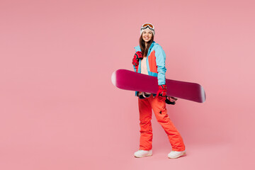 Poster - Full body side view young smiling woman wear warm blue padded jacket ski goggles mask spend weekend winter season in mountains hold snowboard walk go isolated on plain pink background. Hobby concept.