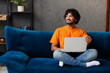 © ViDi Studio - Young IT Indian man wear orange casual clothes sit on blue sofa couch hold use work on laptop pc computer stay home hotel flat rest relax spend free spare time in living room indoor. Lounge concept.