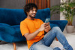 © ViDi Studio - Side view young Indian man wear orange casual clothes sit near blue sofa couch hold use mobile cell phone stay at home hotel flat rest relax spend free spare time in living room indoor Lounge concept