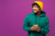 © ViDi Studio - Young happy Indian man wear green hoody yellow hat casual clothes hold in hand use mobile cell phone look aside on area mock up isolated on plain purple background studio portrait. Lifestyle concept.