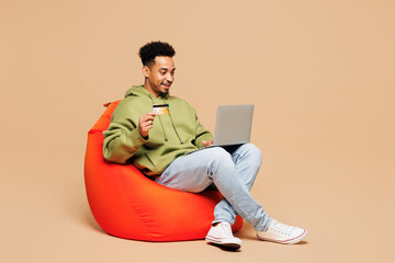 Wall Mural - Full body young IT man of African American ethnicity wear green hoodie casual clothes work hold use laptop pc computer credit bank card shopping online isolated on plain pastel light beige background