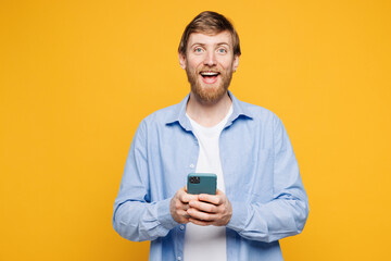 Wall Mural - Young shocked surprised happy Caucasian man wear blue shirt t-shirt casual clothes hold in hand use mobile cell phone look camera isolated on plain yellow background studio portrait Lifestyle concept