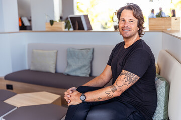 A man with tattoos on his arm sits on a couch smiling in a room