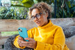 © simona - Attractive woman with curly hair wearing glassesholding a smartphone and scrolling social media apps in a relaxed mood during a quiet moment outdoors.  Wear a yellow sweater, sitting alone in garden,