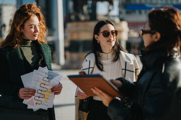 Wall Mural - Three businesswomen engage in an outdoor discussion about project plans and data analysis charts. They share documents and collaborate in a lively, professional atmosphere in a sunny urban setting.