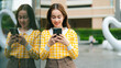 © Leny Studio - young woman in yellow checkered shirt smiles while using her smartphone, standing near reflective glass wall in urban outdoor setting