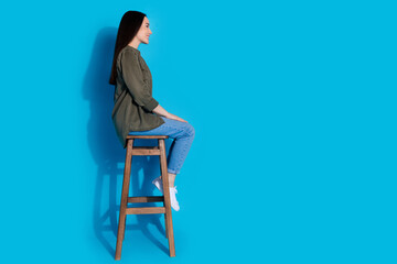 Wall Mural - Young woman sitting on a wooden stool against a vibrant blue background, wearing casual attire