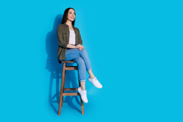 Wall Mural - Young woman seated on a stool against a vibrant blue background, wearing casual clothes and smiling