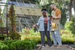 © Seventyfour - Full length of professional African American landscaper pointing at flower bed with decorative conifer shrubs planning garden project on tablet computer with male worker in plant nursery, copy space