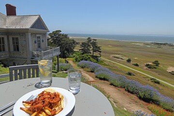 A scenic outdoor dining setup featuring a plate of pasta and fries, with a breathtaking view of a coastline and vibrant flowers.