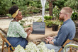 © Seventyfour - Side view of professional female landscape architect of Black ethnicity sitting at garden table with laptop displaying calendar page for planning, talking to male client about design project outdoors