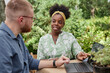 © Seventyfour - Positive Black female landscape designer pointing at laptop screen discussing garden project with male client while sitting at table in sunlit garden