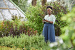 © Seventyfour - Full length of African American female landscape designer using mobile phone while waiting for meeting with client in green lively alley of plant nursery, copy space