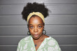 © Seventyfour - Portrait shot of African American female designer wearing transparent earrings and wide textile headband confidently looking at camera while posing against grey garage door, camera flash, copy space