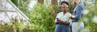 © Seventyfour - Website header shot of positive African American female landscape designer using phone while discussing with male client garden project captured against lively green trees in plant nursery, copy space