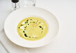 © Food Images - A bowl of smooth yellow soup is elegantly presented with a drizzle of green oil. The setting includes a clear glass of water and a salt grinder, creating a refined dining experience