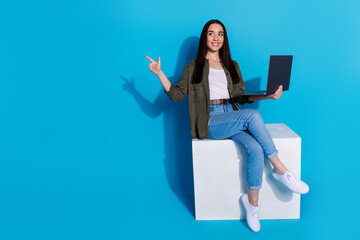 Wall Mural - Stylish woman with laptop gesturing, sitting on a white cube against blue background, showing confident and positive energy