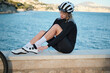 © KatyaPulka - Female cyclist rests by the sea, adjusting her cycling shoes.  She wears a black cycling kit and white cycling shoes. The ocean provides a stunning backdrop.