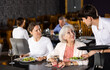 © JackF - Young guy waiter in uniform serves ordered dish to elderly woman and adult woman in restaurant