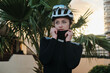 © KatyaPulka - Young woman cyclist adjusts her neck warmer while wearing a helmet and sunglasses. Palm trees and city buildings in the background. Perfect for active lifestyle, sports, and outdoor themes.