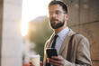 © Zamrznuti tonovi - Serious bearded businessman using smartphone and holding coffee outside office building