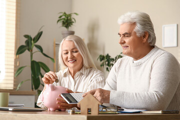 Wall Mural - Senior couple with calculator putting coin into piggy bank at table in kitchen