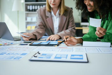 An African American businesswoman and a Caucasian woman collaborate at a desk, brainstorming wireframe interface designs for a mobile app, discussing UI/UX, customer descriptions,color codes enhance