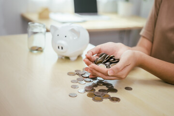 A young woman saves money for the future by putting coins in a piggy bank on the table, managing monthly expenses, and focusing on investment and business growth for financial stability