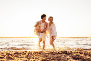 Wall Mural - Smiling beautiful woman and her handsome boyfriend. Excited couple in casual clothes. Happy cheerful family. Female and man run at sunrise over sea beach outdoors. Run at seaside in summer day