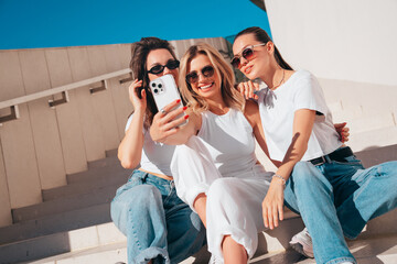 Wall Mural - Three young beautiful smiling hipster female in trendy summer white t-shirt  and jeans clothes. Sexy carefree women posing in street. Positive models having fun, going crazy. In sunny day, take selfie