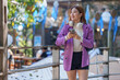 © crizzystudio - Young asian freelancer, wearing a purple blazer and headphones, smiles while using her smartphone in an urban setting, leaning against a railing