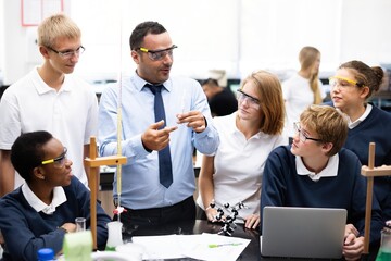 Fototapete - Diverse students and teacher in a science lab. Group of students learning chemistry. Teacher explaining experiment. Students wearing safety goggles. Teacher teaching a diverse high school class.
