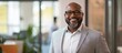 © StockKing - Smiling African American businessman in a light gray suit standing confidently in a modern office with greenery and large windows in background.