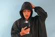 © Lailar Studio - Asian man wearing raincoat holding cell phone with confused expression