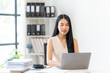 © Wicitr - Focused on the Task: A young Asian businesswoman with a confident smile, diligently works on her laptop at her office desk. The image conveys focus, determination, and professionalism.