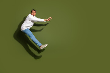 Young man in a stylish casual white sweater and blue jeans jumps playfully against a vibrant khaki background