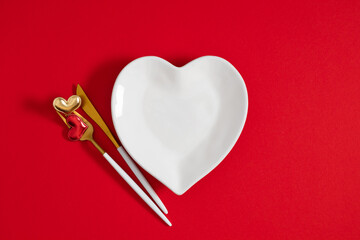 Wall Mural - White empty heart-shaped plate and cutlery fork and knife, hearts valentine on red background. Holiday dinner to celebrate Valentine's Day, concept