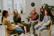 © AnnaStills - Group of women seated in circle discussing personal topics in cozy therapy room surrounded by plants and light from large windows. Sharing experiences and support in comfortable environment