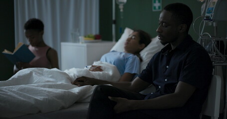 Wall Mural - Young African mother sits in a hospital room next to her unconscious child, reading a book, while her father waits for the child to wake up.