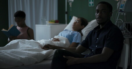 Wall Mural - Young African mother sits in a hospital room next to her unconscious child, reading a book, while her father waits for the child to wake up.