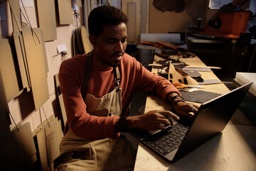 Poster - Man engaged in leather crafting project in a dimly lit workshop, utilizing laptop for design reference and planning