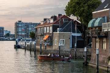 Dordrecht, South Holland, Holland - 09.07.2024: A scenic canal with a wooden boat carrying passengers. Historic brick houses with colorful awnings line the waterfront. Green trees and modern buildings