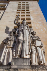 Wall Mural - A stone sculpture group featuring three figures with symbolic objects stands against a textured building facade under a bright blue sky.