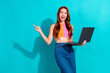 © deagreez - Energetic young woman with red hair pointing and holding a laptop against a bright teal background in summer casual outfit and flared jeans