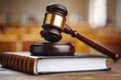 © Yuliia - close-up shot of a golden gavel resting on top of a thick law book with a blurred background of a wooden courtroom table, representing the concept of justice and legal proceedings.